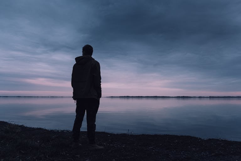 A solitary silhouette of a man in a jacket gazing at a lake during a peaceful sunset, creating a serene atmosphere.