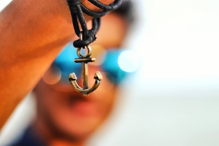 Close-up of a hand holding an anchor pendant, blurred face and sunglasses in background.